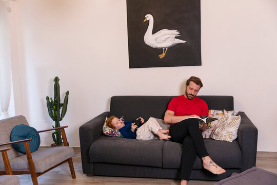 Man reading magazine while son playing with joystick on sofa at home