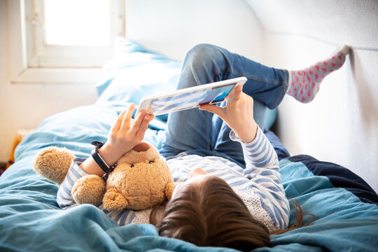 Little girl lying on bed with teddy bear using digital tablet
