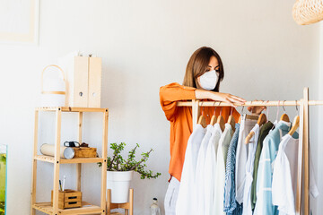 Female fashion designer working at home with clothes stand wearing protective face mask