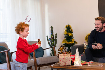 Smiling father looking at cute son wearing antler's headband playing with toy in living room