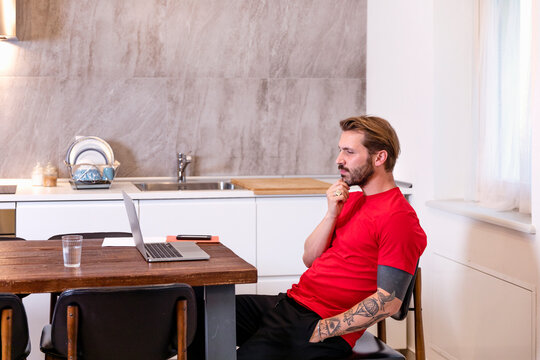 Thoughtful mid adult man looking at laptop on dining table while sitting in kitchen