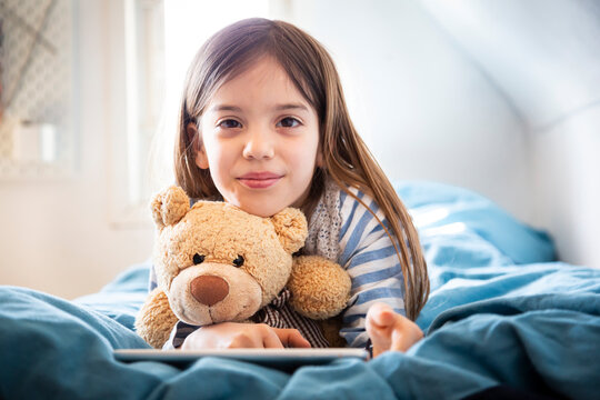 Portrait of smiling girl lying on bed with teddy bear and digital tablet