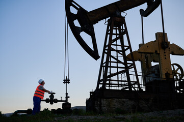 Petroleum operator in work vest using pumping unit to extract crude oil from oil well. Oilman...