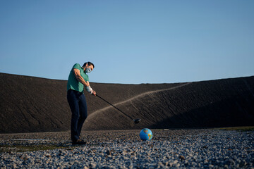 Mature man with a mask playing golf with a globe on a disused mine tip