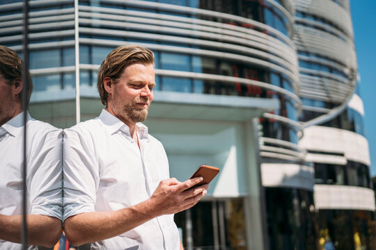 Businessman in the city leaning against glass front checking smartphone