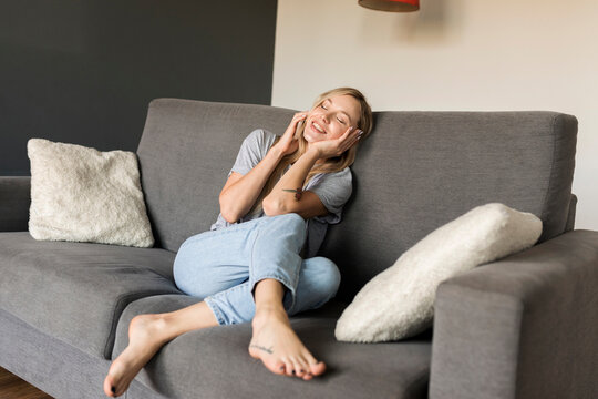 Smiling young woman with closed eyes sitting on couch talking on cell phone
