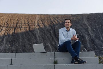 Mature businessman sitting on stairs on a disused mine tip next to laptop