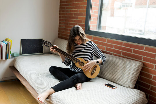 Young woman sitting on couch at home playing guitar