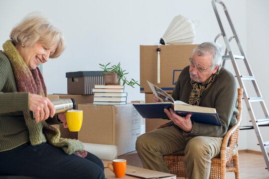 Senior couple with photo album and hot drink surrounded by cardboard boxes in an empty room