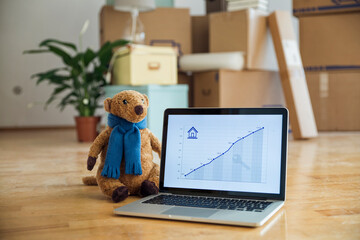Teddy bear and rising line graph on laptop screen in front of cardboard boxes in a new home