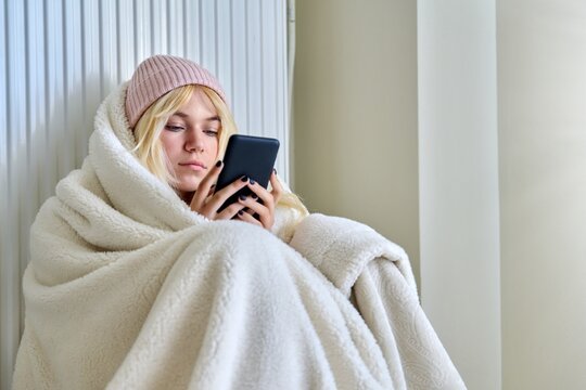 Cold season, frozen teenager in hat under blanket sitting near a heating radiator with smartphone