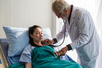 Doctor and patient in the patient room at hospital. Senior male doctor using stethoscope examining woman patient’s heartbeat. Male doctor visiting and checking female patient. Health care concept