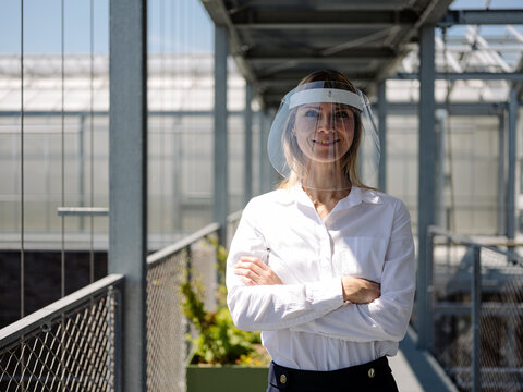 Smiling businesswoman wearing face shield with arms crossed standing in greenhouse