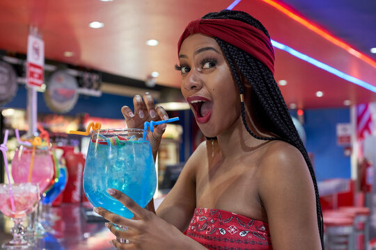 Young woman with braided hairstyle sitting on a restaurant's bar, drinking a blue cocktail