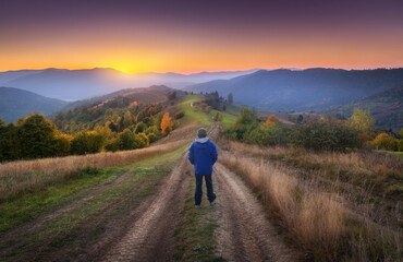 Obraz premium Man on the rural dirt road on the hill looking on mountains in fog at colorful sunset in autumn in Ukraine. Landscape with guy, foggy hills, green grass, meadows, trail, forest, orange sky in fall