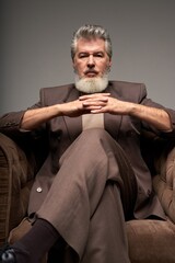 Portrait of serious mature businessman with beard wearing elegant suit looking at camera, sitting with hands crossed in armchair while posing isolated over gray background