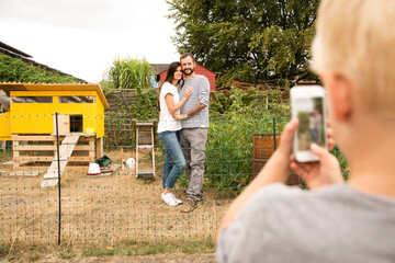 Boy taking cell phone picture of parents standing at chickenhouse in garden