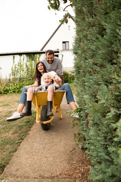Playful man pushing wife and son sitting in wheelbarrow in garden