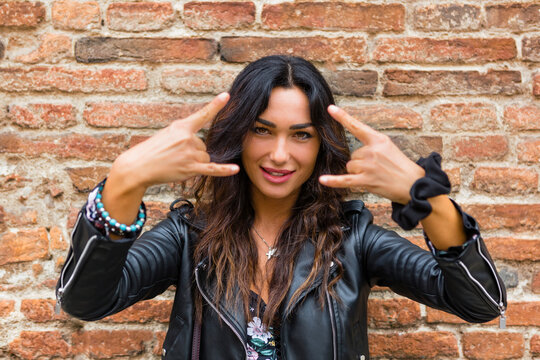 Portrait of young woman wearing black leather jacket and showing rock and roll sign, brick wall