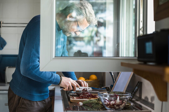 Mature man preparing salad at open window in his kitchen