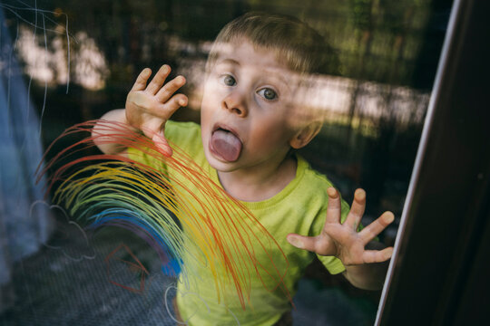 Portrait of little boy standing behind balcony door licking glass pane with drawn rainbow