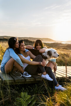 Three happy women with dog sitting on boardwalk in dunes at sunset