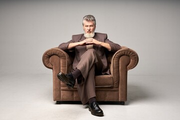 Full length shot of brutal mature businessman with beard wearing elegant suit looking at camera, sitting with hands crossed in armchair while posing isolated over light background - Powered by Adobe