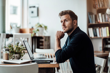 Mid adult businessman looking away while sitting by laptop at cafe
