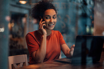 Smiling woman with laptop talking on mobile phone while sitting at cafe