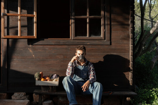 Male farmer with vegetable crate having coffee against wooden house on sunny day