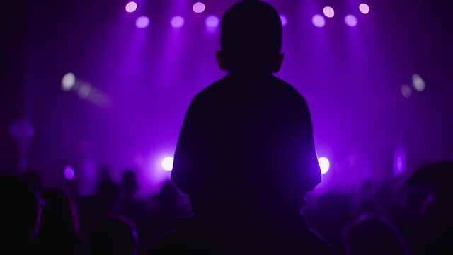 silhouette fan kid boy on father shoulders enjoying listening to artist during concert at night, people on background of stage with lights in purple color