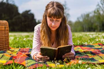 Young woman lying on a picnic blanket, reading a book in a park