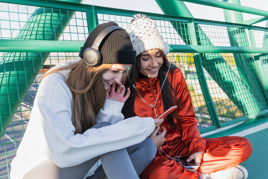 Smiling friends wearing headphones using mobile phone while sitting on bridge