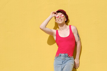 Young non-binary model smiling while posing outdoors leaning against a yellow wall.