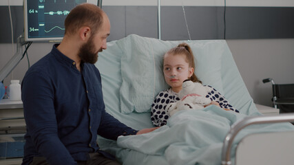 Father sitting beside sick daughter discussing sickness therapy explaining medication treatment during disease examination in hospital ward. Little kid lying in bed after suffering medical surgery