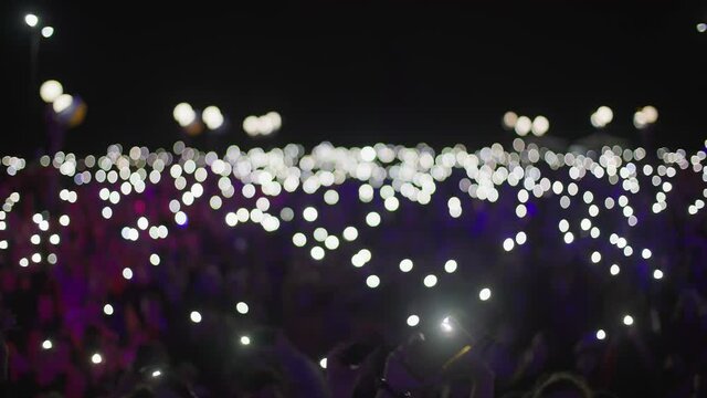 Unrecognizable Group Of Fans Holding Cell Phones With Flashlights In Theirs Hands In Night Time, Atmosphere Of Concert Out Of Focus