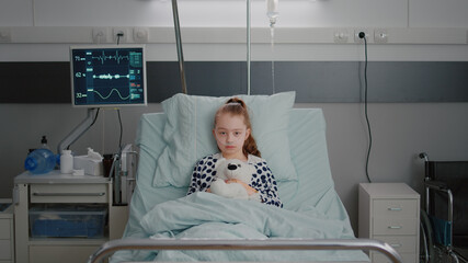 Portrait of hospitalized sick girl child patient holding teddy bear resting in bed during medical...