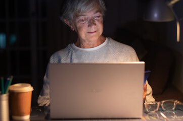 Elderly woman browsing social media content using laptop on desk holding mobile phone. Old retired...