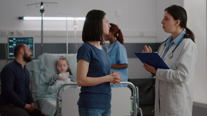 Pediatrician woman doctor explaining recovery treatment to worried mother while black nurse monitoring sickness symptoms. Sick child resting in bed recovering after disease breathing surgery