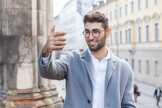 Germany, Munich, portrait of young businessman taking selfie with cell phone in the city
