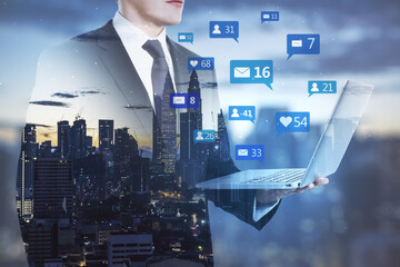 Obraz premium Businessman holding laptop with social media icons on blurry night city background. Communication, network, connections, device and app concept. Double exposure.