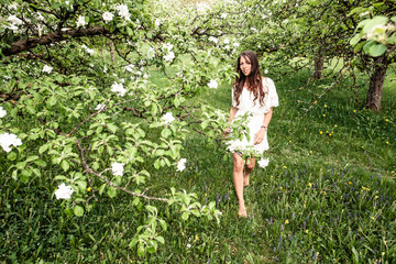 Young woman wearing white dress walking barefoot in garden with blossoming apple trees