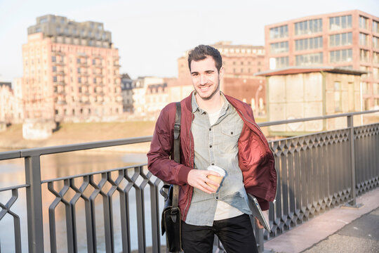 Smiling young man with tablet, bag and takeaway coffee on a bridge in the city
