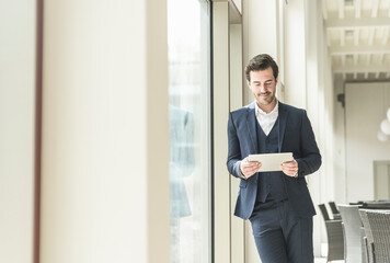 Young businessman standing in office building, using digital tablet, leaning on window