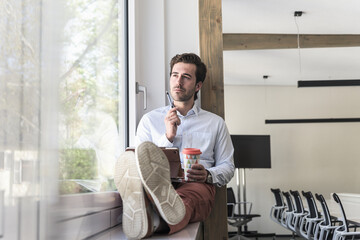 Young businessman sitting on windowsill, using digital tablet, drinking coffee