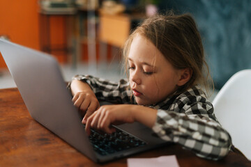 Close-up side view of pretty elementary child girl using typing on keyboard of laptop sitting at desk in cozy living room. Cute little primary serious pupil schoolgirl e-learning online using computer