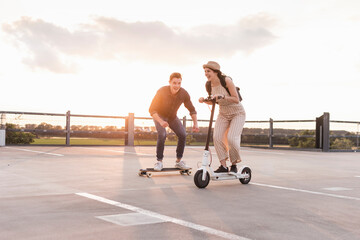 Young man and woman riding on longboard and electric scooter on parking deck at sunset