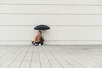 Young man with umbrella, sitting on ground, looking uncertain