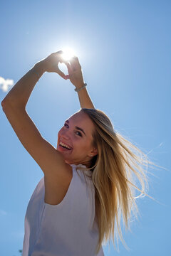 Excited young woman making heart shape with hands against sky in sunlight