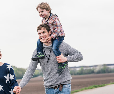 Happy family walking together next to fields, father carrying son piggyback